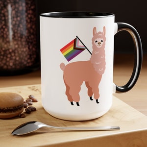 May include: White ceramic mug with a black rim and handle. The mug features a cartoon illustration of a llama holding a rainbow pride flag.