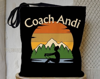May include: Black tote bag with a retro sunset graphic and the text "Coach Andi". The graphic features mountains, trees, and a silhouette of a person rowing a boat.