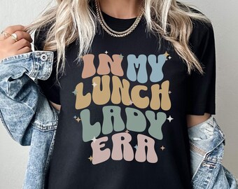 May include: Black t-shirt with the text "In My Lunch Lady Era" in colorful retro font.