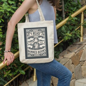 May include: A beige tote bag featuring a black and white graphic of a cat reading a book. The graphic reads "I Read Banned Books" and "Keep Your Paws Off Our Books".