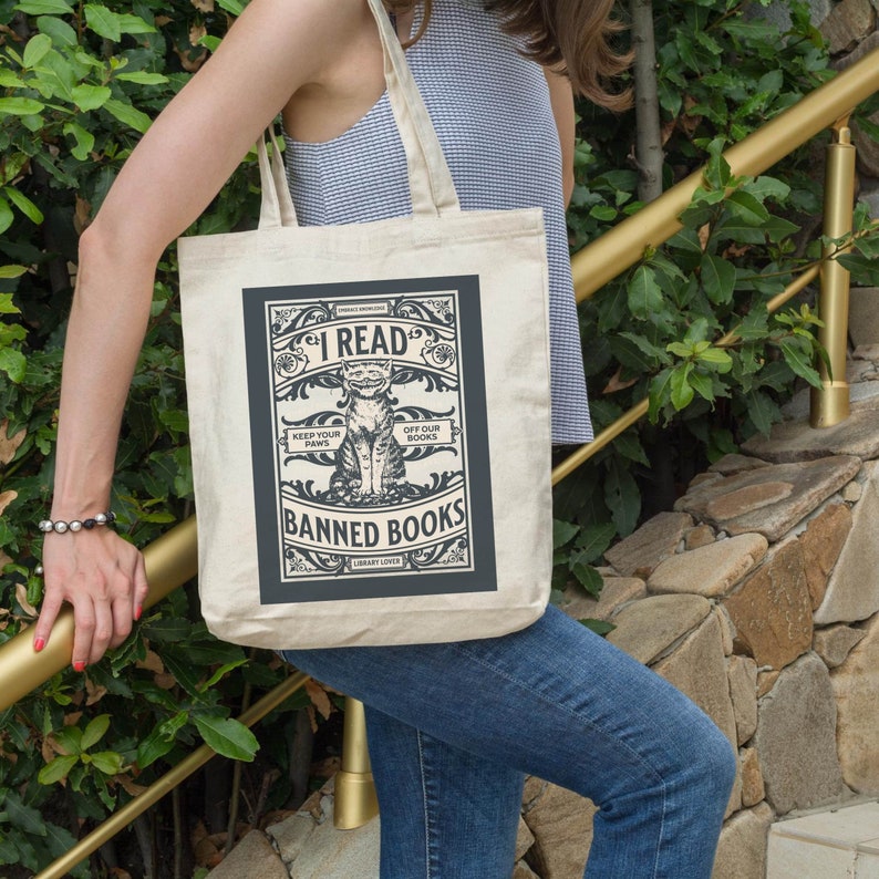 May include: A beige tote bag featuring a black and white graphic of a cat reading a book. The graphic reads "I Read Banned Books" and "Keep Your Paws Off Our Books".