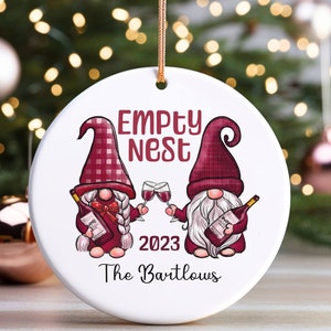 May include: White round Christmas ornament with two red gnomes holding wine glasses and bottles of wine. The ornament says "Empty Nest 2023 The Bartlows".