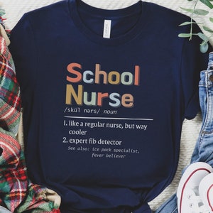 Peut inclure: Un t-shirt bleu marine avec le texte "School Nurse" en lettres color&eacute;es. Le texte est d&eacute;fini comme "comme une infirmi&egrave;re ordinaire, mais bien plus cool" et "d&eacute;tecteur de mensonges expert".