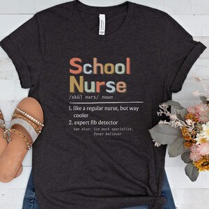 Peut inclure: Un t-shirt gris fonc&eacute; avec le texte "School Nurse" en lettres color&eacute;es. Le texte est d&eacute;fini comme "comme une infirmi&egrave;re ordinaire, mais bien plus cool" et "d&eacute;tecteur de mensonges expert".