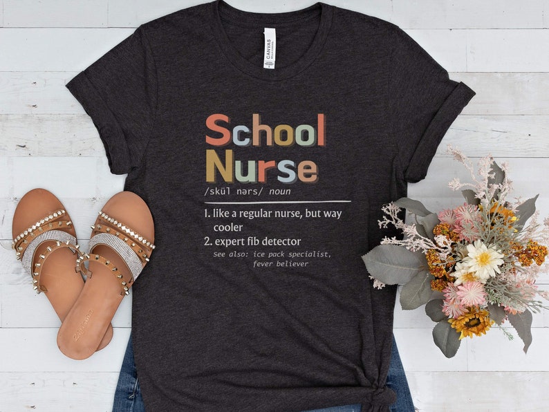 Peut inclure: Un t-shirt gris fonc&eacute; avec le texte "School Nurse" en lettres color&eacute;es. Le texte est d&eacute;fini comme "comme une infirmi&egrave;re ordinaire, mais bien plus cool" et "d&eacute;tecteur de mensonges expert".