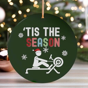May include: Green round Christmas ornament with the text "'Tis the season" in white letters with a red, green, yellow, and blue outline. A white silhouette of a person rowing a rowing machine is in the bottom half of the ornament.