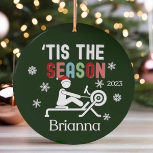 May include: Green round Christmas ornament with a white stick figure rowing on a rowing machine. The ornament has the text 'Tis the Season' in colorful letters and the year '2023' in white. The name 'Brianna' is printed at the bottom of the ornament.