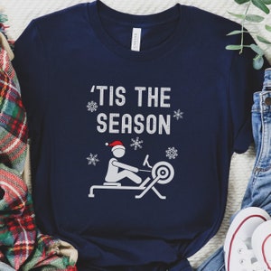 May include: Navy blue t-shirt with a white graphic of a person rowing a rowing machine with the text 'Tis the season' above the graphic.