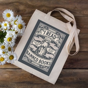 May include: A white tote bag with a blue design featuring a cat and the text "I Read Banned Books". The design is in a vintage style with ornate lettering and illustrations.
