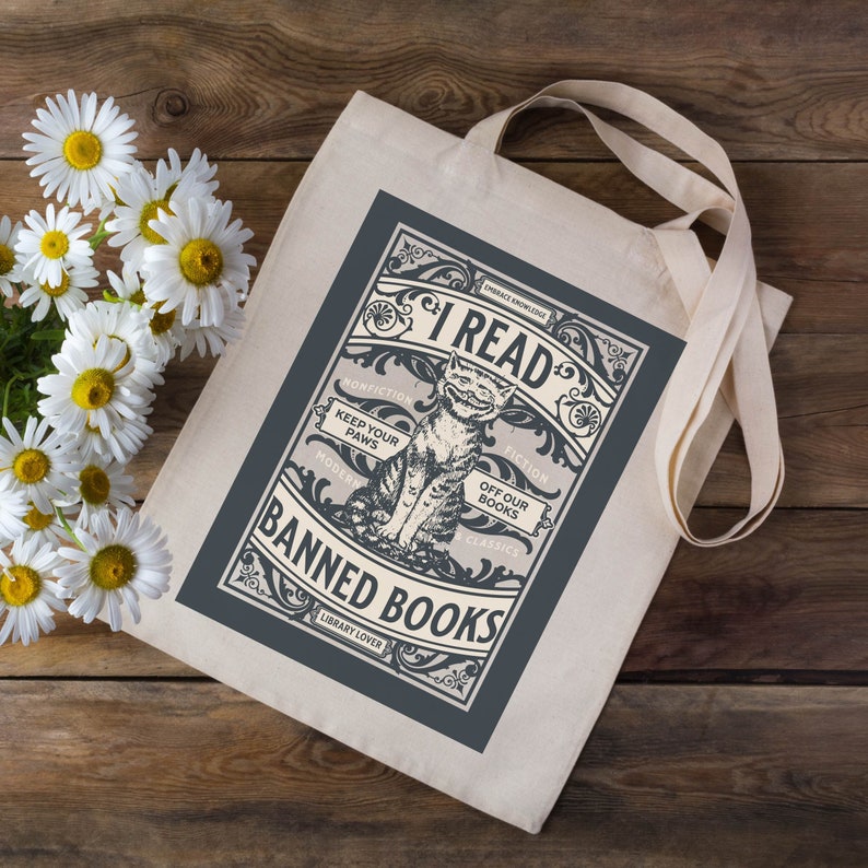 May include: A white tote bag with a blue design featuring a cat and the text "I Read Banned Books". The design is in a vintage style with ornate lettering and illustrations.
