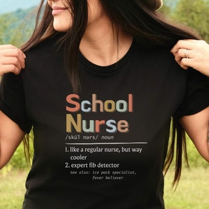 Peut inclure: Un t-shirt noir avec le texte "School Nurse" en lettres color&eacute;es. Le texte est d&eacute;fini comme "comme une infirmi&egrave;re ordinaire, mais bien plus cool" et "d&eacute;tecteur de mensonges expert".