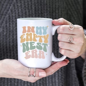 May include: White ceramic mug with the text "In My Empty Nest Era" in colorful, retro-style lettering. The mug is being held by a person's hands.