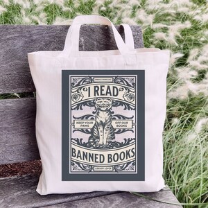 May include: A white tote bag with a grey design featuring a cat with a mischievous grin. The design says "I Read Banned Books" with the text "Embrace Knowledge" and "Library Lover" at the top and bottom of the design. The cat is sitting on a book with the text "Keep Your Paws Off Our Books" on the book.