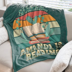 A large dark teal green throw blanket is spread over a sofa. It pictures a stack of book with some leaves and the text, Shhhhh...Amanda is reading. Behind the stack of books is a circular sunset graphic in colors ranging from red to teal.