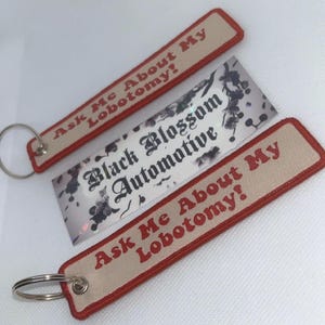 Puede incluir: Tres llaveros. Dos tienen el texto "Ask Me About My Lobotomy!" en rojo sobre un fondo beige con un borde rojo. Se adjunta un llavero plateado. También hay una tarjeta de visita con "Black Blossom Automotive".
