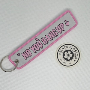 Puede incluir: Llavero rosa y blanco con un corazón rosa y el texto "NO YOU HANG UP" en una fuente que gotea. El llavero está unido a un llavero plateado. Una pegatina en blanco y negro con una flor y el texto "BLACK BLOSSOM AUTOMOTIVE" está en el fondo.