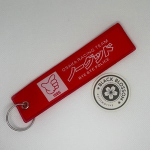 Könnte beinhalten: Roter Schlüsselanhänger mit einem weißen Daumen-hoch-Symbol und dem Text "OSAKA RACING TEAM ノックアウト BYE BYE POLICE 1985". Der Schlüsselanhänger hat auch einen schwarz-weißen Aufkleber mit dem Text "BLACK BLOSSOM AUTOMOTIVE" und einer stilisierten Blume.
