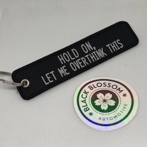 Puede incluir: Llavero negro con una frase bordada en blanco "Hold on, let me overthink this". El llavero está unido a un llavero plateado. Una pegatina holográfica con el texto "Black Blossom Automotive" está en el fondo.