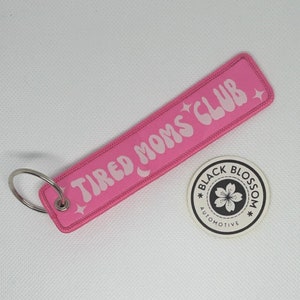 Puede incluir: Llavero rosa con el texto "TIRED MOMS CLUB" en blanco y estrellas. El llavero está unido a un anillo plateado. Un pequeño círculo blanco con una flor negra y el texto "BLACK BLOSSOM AUTOMOTIVE" está en el fondo.