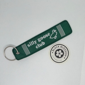 May include: Green fabric keychain with a white goose and the text "silly goose club". The keychain is attached to a silver key ring. A white sticker with a black flower and the text "Black Blossom Automotive" is in the background.
