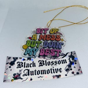 Puede incluir: Un adorno de acrílico colorido con el texto "Bit of a mess but doin' my best". El adorno está sujeto a una cuerda dorada y tiene una pegatina con el texto "Black Blossom Automotive" en la parte inferior.