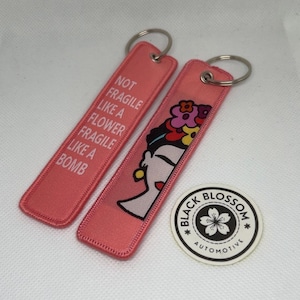 Puede incluir: Dos llaveros rosas con aros plateados. Uno tiene el texto "NOT FRAGILE LIKE A FLOWER FRAGILE LIKE A BOMB" en blanco. El otro presenta una colorida ilustración del perfil de una mujer. También hay una pegatina redonda con el texto "BLACK BLOSSOM AUTOMOTIVE".