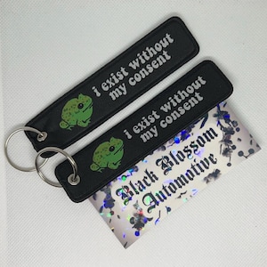 May include: Two black keychains with a green frog graphic and the text "i exist without my consent". The keychains are attached to a silver keyring. The keychains are on top of a holographic card with the text "Black Blossom Automotive".