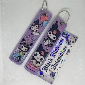 Può includere: Due portachiavi viola con personaggi dei cartoni animati in bianco e nero. I portachiavi sono attaccati a anelli portachiavi argentati. I portachiavi sono su una superficie bianca con un adesivo olografico che dice "Black Blossom Automotive".