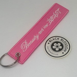 Könnte beinhalten: Rosafarbener Schlüsselanhänger mit weißer Stickerei, auf der "Beauty and Her Beast" steht. Der Schlüsselanhänger hat einen silbernen Ring und ein weißes rundes Etikett mit einer schwarzen Kirschblüte und dem Text "Black Blossom Automotive".