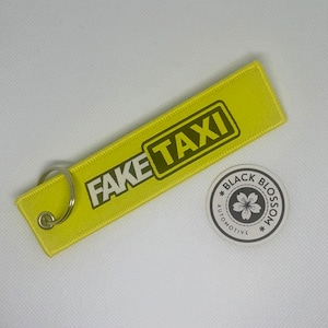 May include: Yellow key chain with the text "FAKE TAXI" in black and white lettering. The key chain has a silver ring. A small round sticker with the text "BLACK BLOSSOM AUTOMOTIVE" and a stylized flower is in the background.