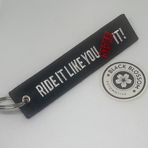 Puede incluir: Llavero negro con bordado blanco que dice "RIDE IT LIKE YOU STOLE IT!" con una línea roja sobre la palabra "STOLE". El llavero está unido a un llavero plateado. Una pegatina circular blanca con una flor negra y el texto "BLACK BLOSSOM AUTOMOTIVE" está en el fondo.