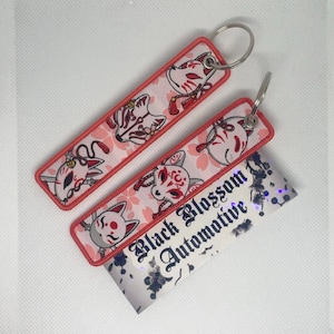 Peut inclure: Deux porte-clés rose et rouge avec un fond blanc représentant un masque de renard japonais. Les porte-clés sont attachés à des anneaux de clés argentés. Les porte-clés sont sur une surface blanche avec un fond noir et blanc qui dit "Black Blossom Automotive".
