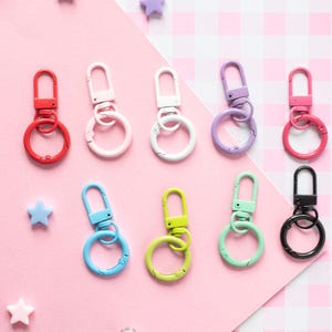 May include: A collection of colorful metal keychains in various colors, including red, pink, white, purple, blue, green, and black. Each keychain features a clasp and a circular ring. The keychains are arranged on a pink and checkered background.
