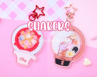May include: Two clear acrylic keychain charms with a shaker design. One charm features a red and white design with the word "Stay" and the other charm features a brown and white design with the word "Atiny".