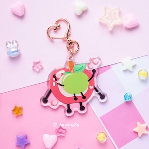May include: A clear acrylic keychain featuring a cartoon apple design with a green center, red outline, and black details. The keychain has a gold-colored heart-shaped clasp. The background is pink with scattered decorative elements.