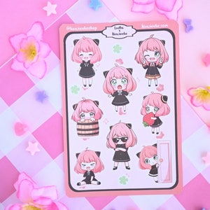 May include: A sheet of pink and white stickers featuring a cartoon character with pink hair and black cat ears. The character is shown in different poses, including holding a strawberry, a barrel, and a door. The text "Ludhe x Kawaii-che" and "kawaiiche.com" is printed on the sheet.