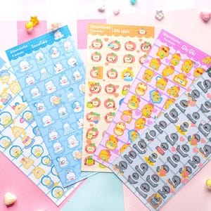 Op de afbeelding: Vijf kleurrijke sticker vellen met diverse cartoon ontwerpen. De vellen tonen karakters zoals sneeuwmannen, appels en luiaards, met teksten als "My star", "Snowflake" en "Little crow". De stickers zijn gerangschikt op een roze en witte achtergrond.