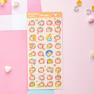 Op de afbeelding: Een stickervel met appelthema-ontwerpen, met schattige gezichten en details. De vellen bevatten de tekst "Little apple" en Japanse tekst. De stickers staan op een lichtcr&egrave;mekleurige achtergrond met een roze en oranje rand.