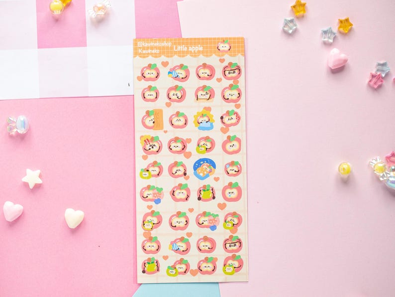 Op de afbeelding: Een stickervel met appelthema-ontwerpen, met schattige gezichten en details. De vellen bevatten de tekst "Little apple" en Japanse tekst. De stickers staan op een lichtcr&egrave;mekleurige achtergrond met een roze en oranje rand.