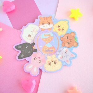 Op de afbeelding: Een set van acht kleurrijke dieren stickers met een kat, een konijn, een hond, een hamster, een vogel, een vos, een beer en een zeemeermin. De stickers zijn in een cirkel rond een zandloper met de woorden "Peace" en "Love" erop gerangschikt.
