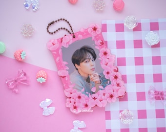 May include: A pink photo frame with a picture of a person, decorated with pink flowers. The frame is surrounded by pink and white decorative elements, including bows, strawberries, and flowers, on a pink background.