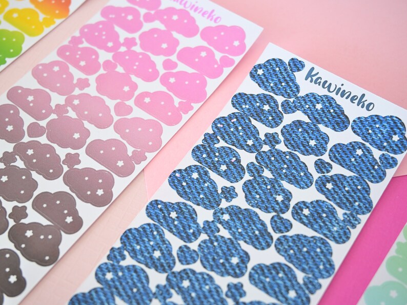 May include: Three sheets of stickers with cloud designs. The first sheet has pink clouds with white stars, the second sheet has gray clouds with white stars, and the third sheet has blue denim-textured clouds with white stars. The text "Kawineko" is printed on the third sheet.