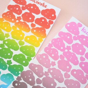 May include: Two sheets of rainbow and pink cloud stickers with white stars. The rainbow sheet has a gradient of red, orange, yellow, green, blue, and purple. The pink sheet has a gradient of light pink to dark pink. Both sheets have the text "Kawineko" printed on them.