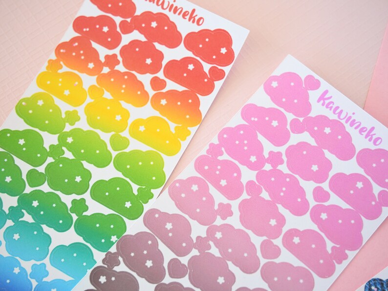 May include: Two sheets of rainbow and pink cloud stickers with white stars. The rainbow sheet has a gradient of red, orange, yellow, green, blue, and purple. The pink sheet has a gradient of light pink to dark pink. Both sheets have the text "Kawineko" printed on them.