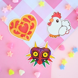 May include: Three colorful stickers on a pink and white checkered background. The stickers are a red heart with gold trim, a white chicken, and a purple, green, and yellow mask with sharp teeth.