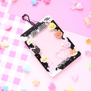 May include: A black and white photo card holder with the text "MY BIAS" and "MY ONLY ONE". The holder is decorated with cartoon animal faces and clouds. The background is pink with scattered star and heart-shaped decorations.