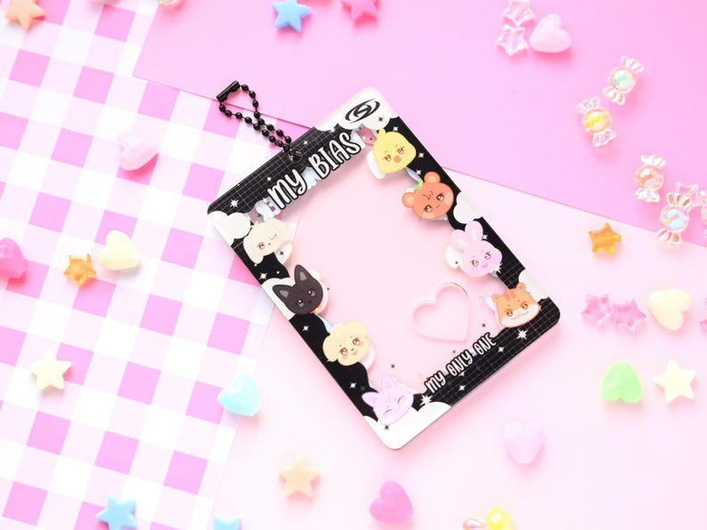 May include: A black and white photo card holder with the text "MY BIAS" and "MY ONLY ONE". The holder is decorated with cartoon animal faces and clouds. The background is pink with scattered star and heart-shaped decorations.