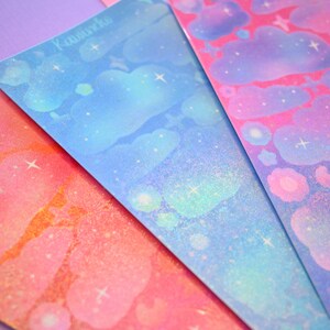 May include: Four sheets of iridescent holographic sticker paper with a cloud and star design. The sheets are in shades of blue, pink, orange, and purple.