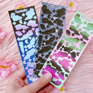 Könnte beinhalten: Drei Blätter mit Aufklebern zum Thema Galaxie. Jedes Blatt zeigt einen Farbverlauf von Rosa, Grün und Blau mit schwarzen Wolken und Sternen. Die Blätter sind mit "Kawineko" beschriftet.