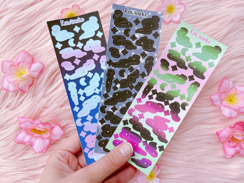 K&ouml;nnte beinhalten: Drei Bl&auml;tter mit Aufklebern zum Thema Galaxie. Jedes Blatt zeigt einen Farbverlauf von Rosa, Gr&uuml;n und Blau mit schwarzen Wolken und Sternen. Die Bl&auml;tter sind mit "Kawineko" beschriftet.